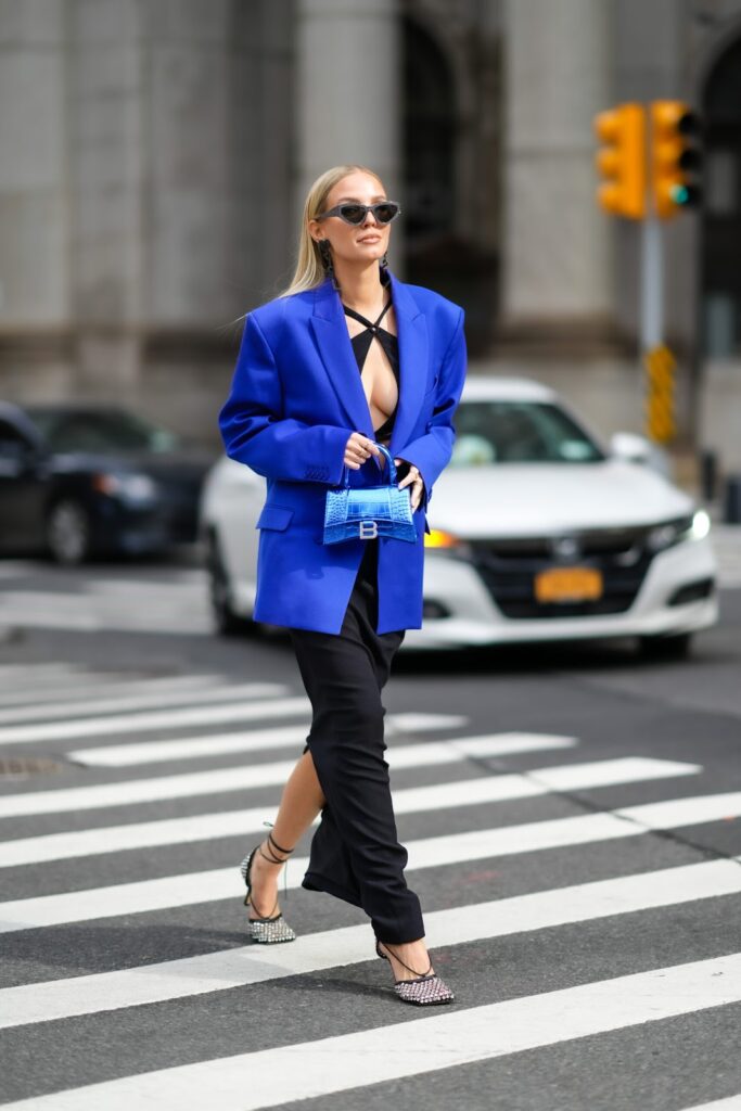 Po ulici kraca zena v modrom saku, ciernych nohaviciach a s modrou kabelkou