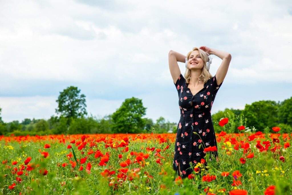 Žena v dlhých čiernych šatách s kvetmi. Stojí v tráve medzi červenými makmi. V ušiach má slúchadlá