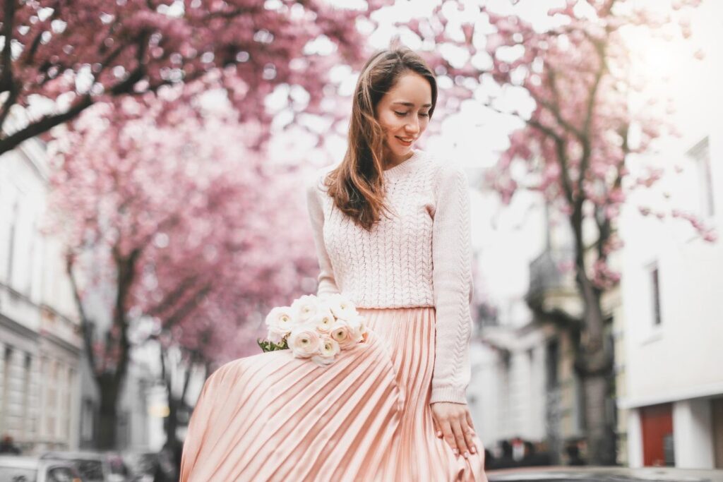 Mladá žena v ružovej plisovanej sukni a ružovom svetri. V ruke drží kyticu kvetov
