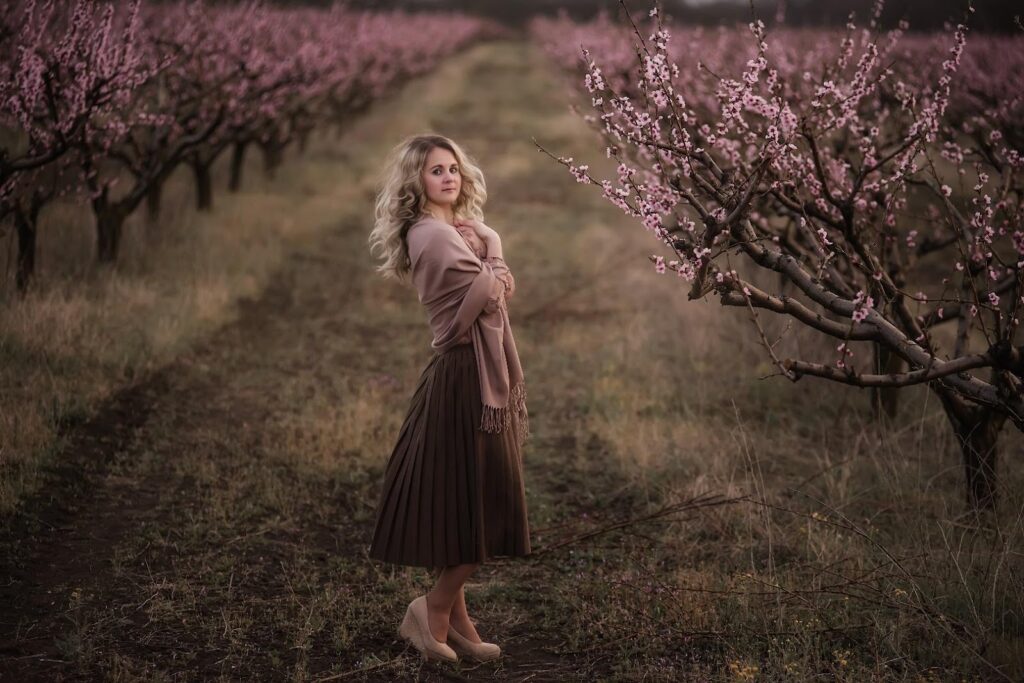 Žena v hnedej plisovanej sukni stojí v sade medzi kvitnúcimi stromami